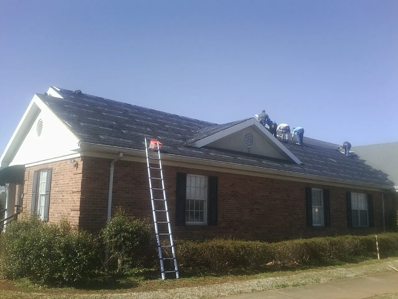 Crew tearing shingles off a brick ranch roof with a ladder set — Emergency Roof Repair work by our crew in Hustle