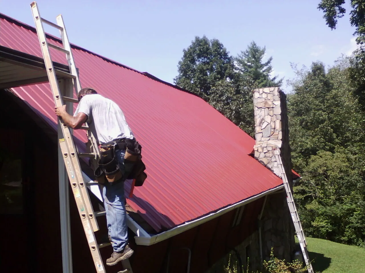 Roofer on a ladder beside a red metal roof and stone chimney — Soffit & Fascia Repair by Roofing Brothers in Hustle